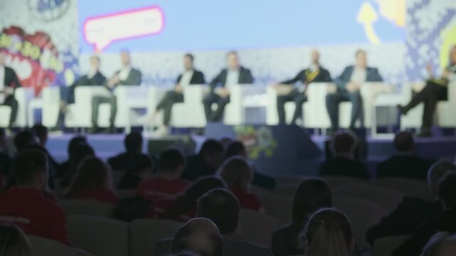 Business leaders sit on stage for panel discussion while audience members listen in dim conference hall.