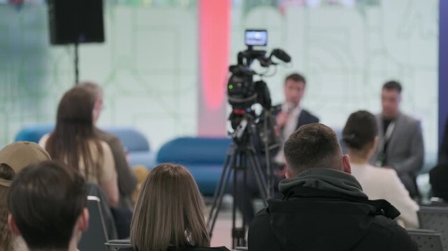 Conference audience listens to panel discussion recorded by professional camera, corporate speakers engage attendees, modern event setup with livestream, media coverage.
