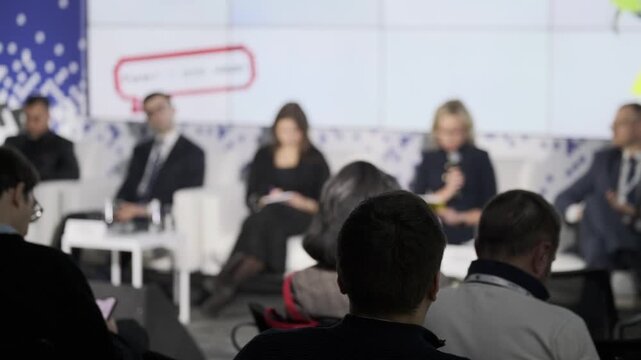 Business conference panel discussion, attendees seated in audience, blurred speakers on stage, professional networking atmosphere.