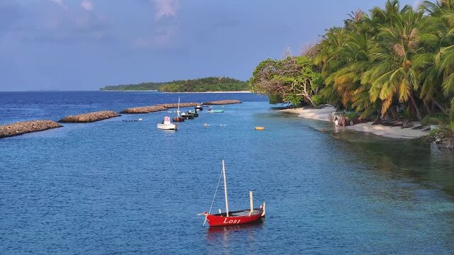 Aerial view of traditional boats anchored in a turquoise lagoon along a lush tropical coastline with palm trees and white sand beaches in Kendhoo, Baa Atoll, Maldives.