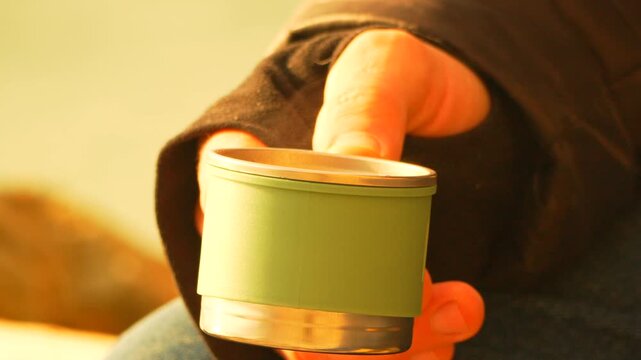Thermos coffee cup hand holding a green travel mug outdoors while steaming hot tea warms a traveler during sunset