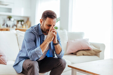 Fototapeta premium Stressed Man Sitting on Sofa Holding Head in Living Room Showing Anxiety and Emotional Exhaustion