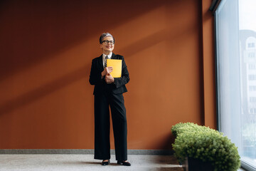 Businesswoman in black suit holding yellow folder stands confidently in modern office with large window and green plants against a warm brown wall © standret