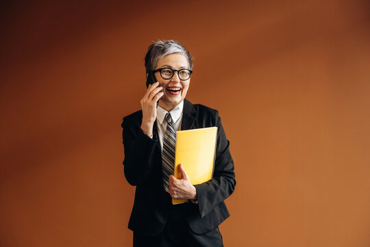Senior Asian businesswoman in black suit holding yellow folder while talking on smartphone, smiling against a warm brown background in a modern office setting