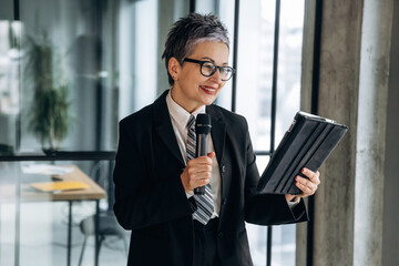 Fototapeta premium Businesswoman in formal attire holding a tablet and microphone while smiling, standing in a modern office with large windows and a contemporary workspace visible