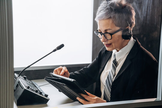 Senior female interpreter wearing headset and glasses uses tablet while seated in soundproof booth with microphone and control panel visible in a modern office setting