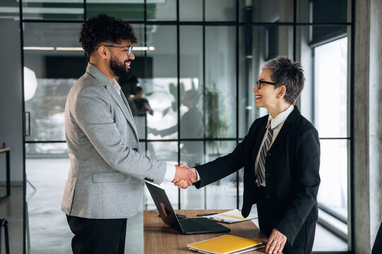 Business professionals shaking hands in a modern office setting, with a laptop and documents on a wooden table, large windows providing natural light