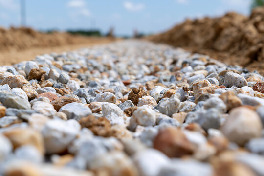 Fresh gravel path stretches toward distant horizon. Blurred dirt mounds frame the foreground. Clear sky with scattered clouds looms above. Construction site under development