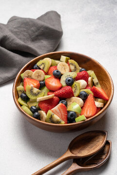 Fresh fruit salad with banana apple kiwi blueberry and strawberry in wooden bowl, top view on light background, healthy eating concept