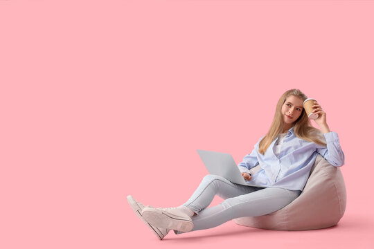 Female programmer with laptop and cup of coffee sitting on pink background