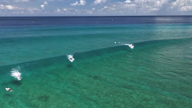 Aerial view of surfers catching a long turquoise wave in the crystal clear Indian Ocean under a blue sky in Thulusdhoo, Kaafu Atoll, Maldives.