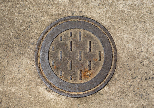 Top-down view of a weathered circular iron manhole cover with rhythmic geometric patterns and rust textures. Minimalist industrial aesthetic featuring rich tactile grit and concrete tones.