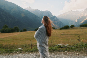 Naklejka premium Young woman wrapped in a gray shawl stands in a windswept meadow, looking toward distant mountains, conveying calm, wonder, and quiet connection with nature