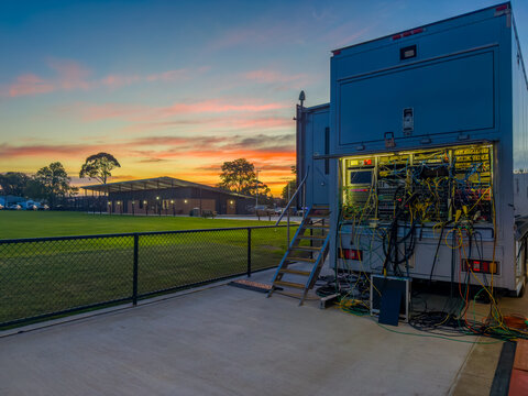 Outside broadcast truck with complex wiring and equipment at a sports stadium during sunset. Outdoor broadcasting van for live television production parked on a concrete platform. Broadcast technology