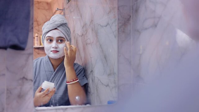 Close up shot of a woman applying facial mask while holding a cosmetic container, standing in front of a bathroom mirror.