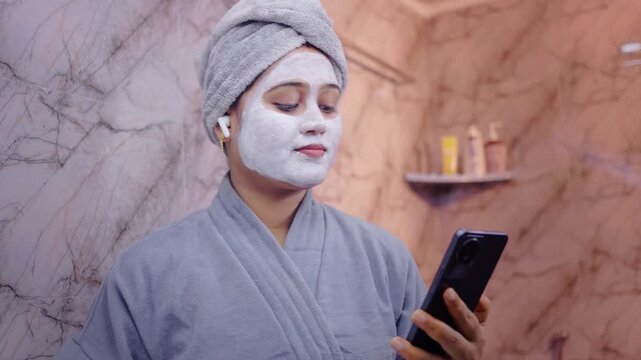 Young lady wearing facial mask while listening to music through earphones and using smartphone in modern bathroom.