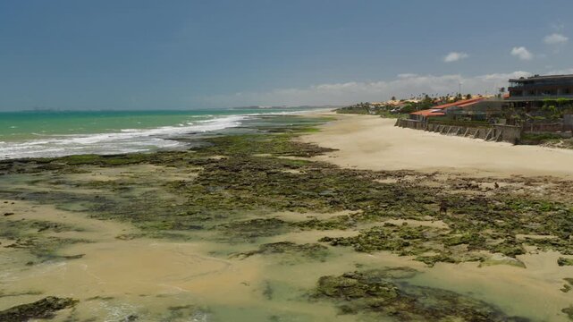 Taiba coastline in Sao Goncalo do Amarante Ceara Brazil South America with rocky intertidal flats waves and beachfront houses on elevated edge showing coastal morphology drone lateral sliding shot