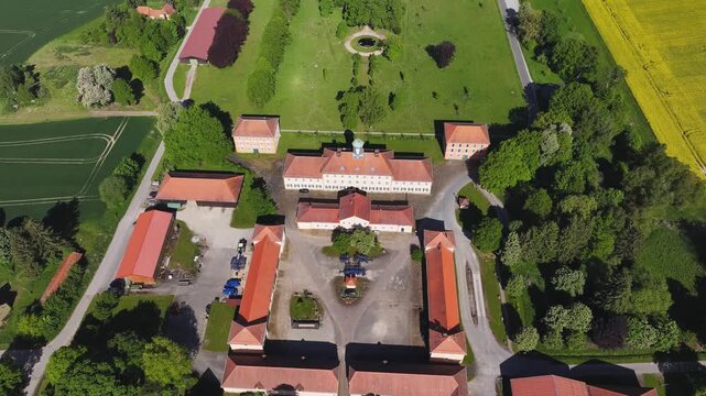 Aerial view of a historic estate with red-roofed buildings, central courtyard, and lush green meadows surrounded by yellow rapeseed fields in Hofgeismar, Hessen, Germany.