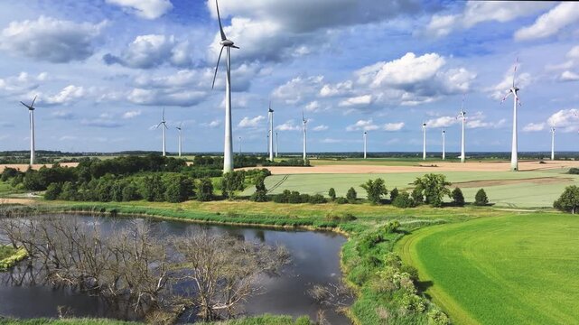 Aerial view of a wind farm with tall turbines standing over green fields and a small pond under a cloudy sky in Uckermark, Brandenburg, Germany.