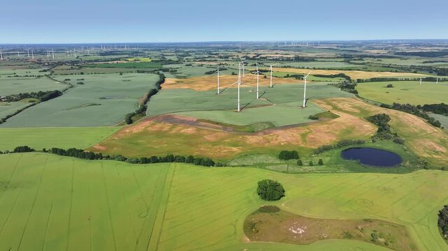 Aerial view of wind turbines standing in vast green and brown agricultural fields with a small pond under a clear blue sky in Uckermark, Brandenburg, Germany.