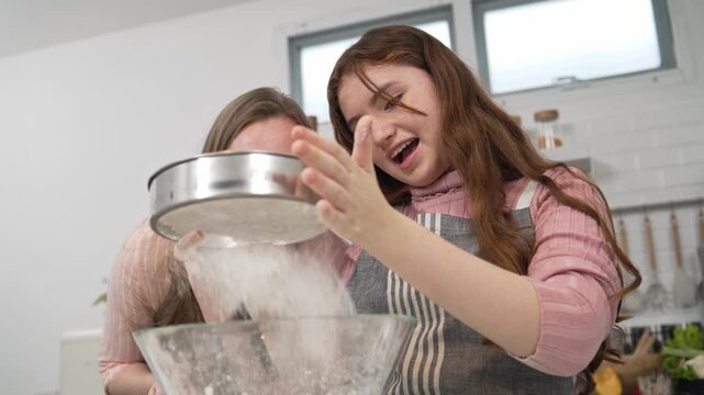 Young girl sifting flour into a glass bowl with her mother in a bright kitchen. Concentrated daughter and mom baking together, preparing ingredients for homemade pastry