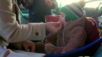 Mother feeding baby in bear teddy fleece coat and striped beanie hat while child sits outdoors in chair, nurturing parent caring for infant during cozy winter family moment © Marco