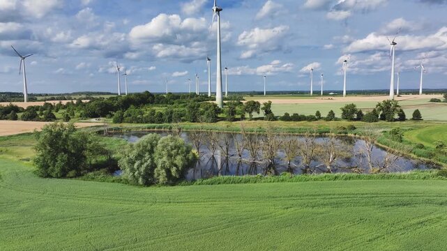 Aerial view of wind turbines on a wind farm near a pond with submerged trees and green fields under a cloudy sky in Uckermark, Brandenburg, Germany.