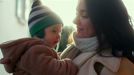 Mother kissing baby in bear teddy fleece coat and striped beanie hat outdoors, woman in beige coat showing affection and warmth to child during cold season family outing © Marco