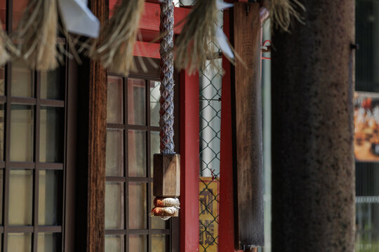 Close-up landscape view of small inner-city shrine illuminated by sliver of sunlight filtering through nearby buildings, spotlighting wooden handle of braided rope hanging from out of frame Suzu bell.
