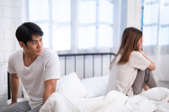 Upset Asian couple sitting apart on bed after argument, relationship problems, emotional distance, marriage conflict and communication issues at home