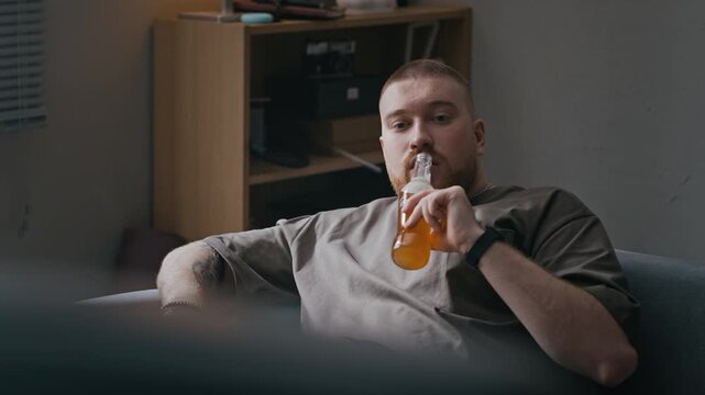 Medium shot of bored Caucasian man with ginger beard relaxing on sofa with beer while watching television in apartment interior