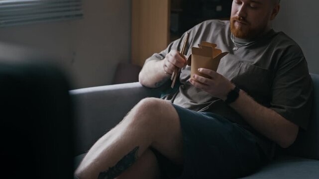 Tilt shot of Caucasian bearded man relaxing on couch with takeout food while wearing ankle monitor in apartment interior