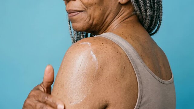 Close-up of a mature African American woman applying moisturizing cream to her shoulder, showing natural skin texture and age spots, dermatology and body care concept