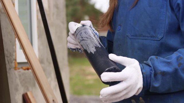 Woman in blue overalls preparing to paint outdoors. Construction worker holding a bottle of black paint, close-up. Worker in blue overalls and white protective gloves holding a plastic can of paint