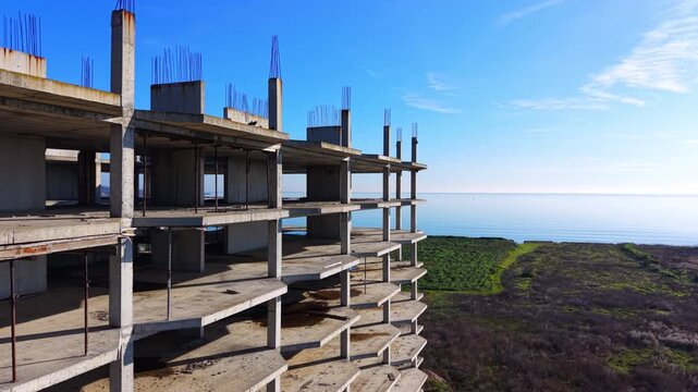Side view of multi story concrete building construction site near ocean under blue sky with green field. Concrete frame of unfinished residential building. Aerial view.
