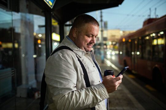 Smiling commuter using smartphone at city bus stop at dusk