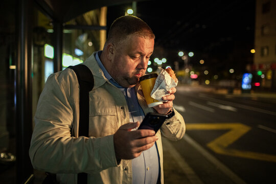 Man using smartphone and drinking coffee at bus stop at night