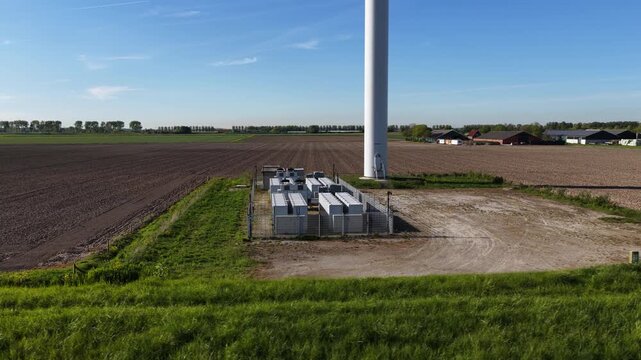 Aerial view of a battery energy storage system at the base of a modern wind turbine, providing a solution for storing clean, sustainable power and ensuring grid stability in a rural landscape