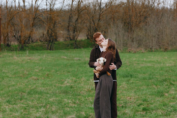 Naklejka premium Young couple embraces tenderly in a green field while holding a small white goat, sharing a romantic moment in casual autumn clothing with trees in the background.