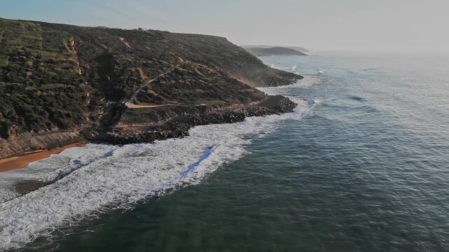 High angle cinematic drone footage of the Atlantic coastline at Praia do Salgado near Nazare, Portugal. Powerful waves and white sea foam wash over the sandy beach during a hazy day