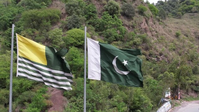 Pakistan and Kashmir flags waving on poles against a green hillside landscape