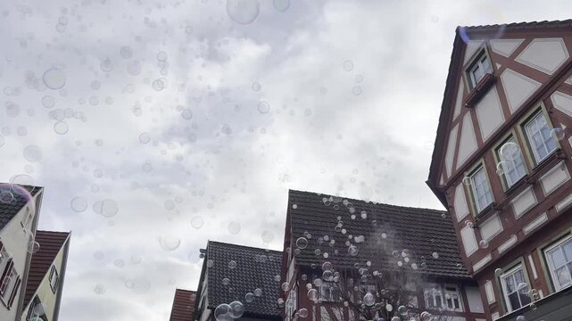 Soap bubbles floating through historic street in Besigheim with traditional half-timbered buildings and cloudy sky. Urban tourism footage with upward camera angle and moving bubbles.