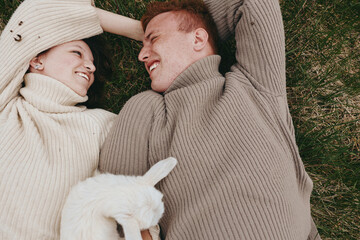 Naklejka premium Joyful couple outdoors, lying on grass with a playful goat kid nearby, cozy sweaters, sunlit scene, relaxed mood, candid affection, natural colors, serene countryside vibe