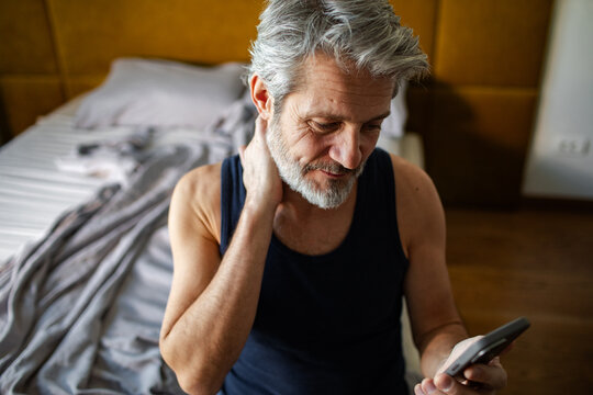 Mature man with neck pain using smartphone in bedroom