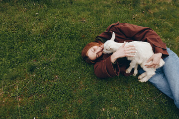 Naklejka premium A woman lies on the grass, hugging a white goat kid with gentle affection. Outdoor scene portraying calm lifestyle, nature, and animal companionship under soft daylight.