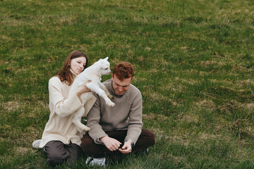 Naklejka premium Couple outdoors with a playful goat kid on a grassy field, capturing a warm moment of affection and connection. Natural light and relaxed mood emphasize lifestyle, care, and rural charm in this