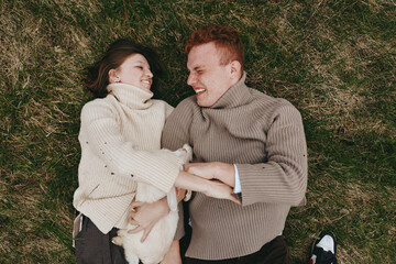 Naklejka premium Cheerful couple outdoors, lying on grass with a baby goat between them, sharing warm smiles, cozy sweaters, natural light, relaxed lifestyle moment in a lush green setting, full of affection