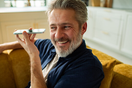 Smiling middle-aged man using smartphone voice assistant at home