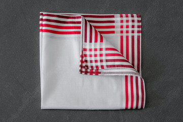 Fototapeta premium A white cotton handkerchief with a bold red striped border and a checkered corner detail, neatly folded on a textured dark background.