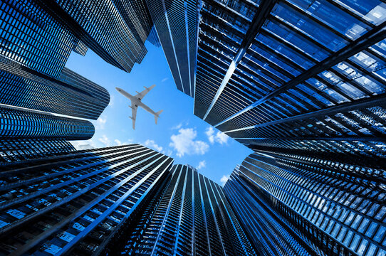Dramatic low angle architectural view of modern skyscrapers with airplane flying overhead against blue sky representing travel aviation business and urban development